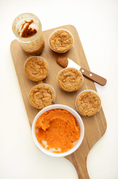 A wooden tray with muffins, almond butter, and a bowl of mashed sweet potato.