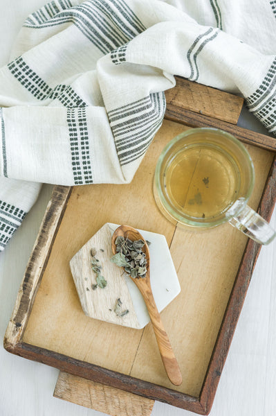 Wooden spoon full of sage tea, a tea cup full of tea, and a textured tea towel on top of a wooden platter.