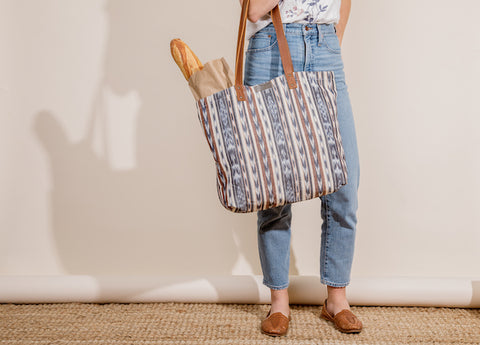 Lower half on women holding farmer's market tote on her forearm with bread and groceries inside. Wearing blue jeans and flat sandals with a white background and tan floor.