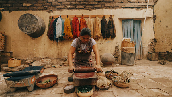 An Artisan working on crushing natural items to create colors that will be used to dye wool. She has all different colors of wool behind her hanging on the wall.