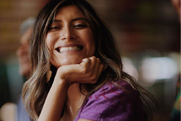 A latin woman sitting at a table smiling at the camera with her hand resting on her chin.