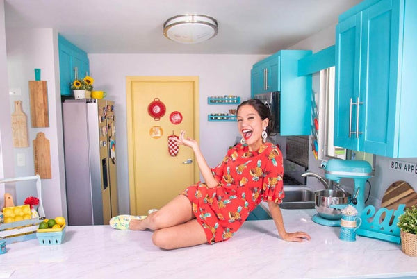 woman in a bright colored kitchen laying on the island holding a cooking tool.