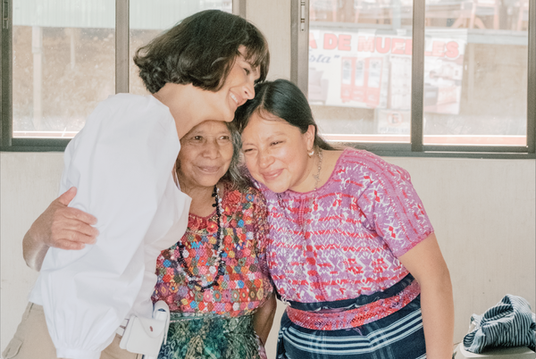 Ali Hynek, CEO fo Nena & Co. hugging two Guatemalan female Artisans in front of some windows inside a house.