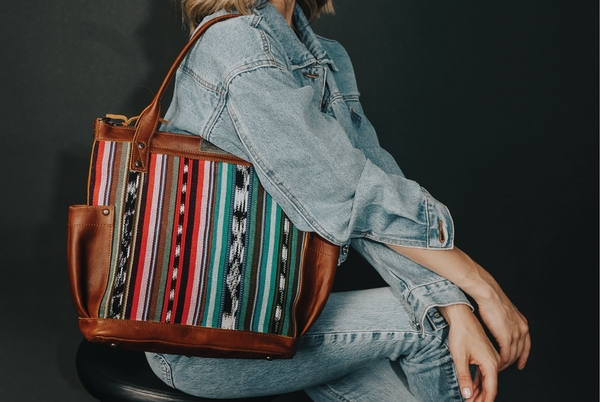 model sitting on a stool sideways with a bag on her shoulder and her arms crossed on her leg.