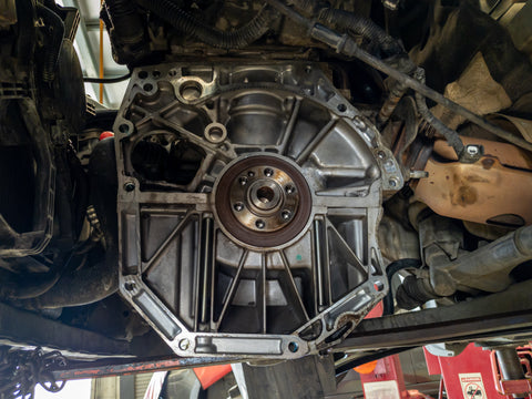 Undercarriage view of a vehicle showing a disassembled engine gearbox with visible flywheel, mounted on a lift in a repair shop