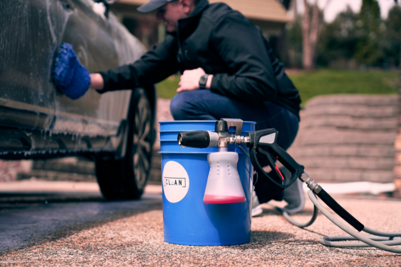 a person sitting on a blue bucket with a spray gun and a car