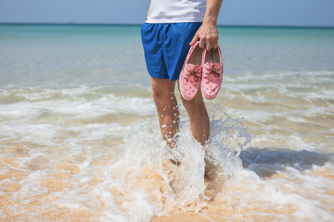man in beach water holding a pair of pink boat shoes
