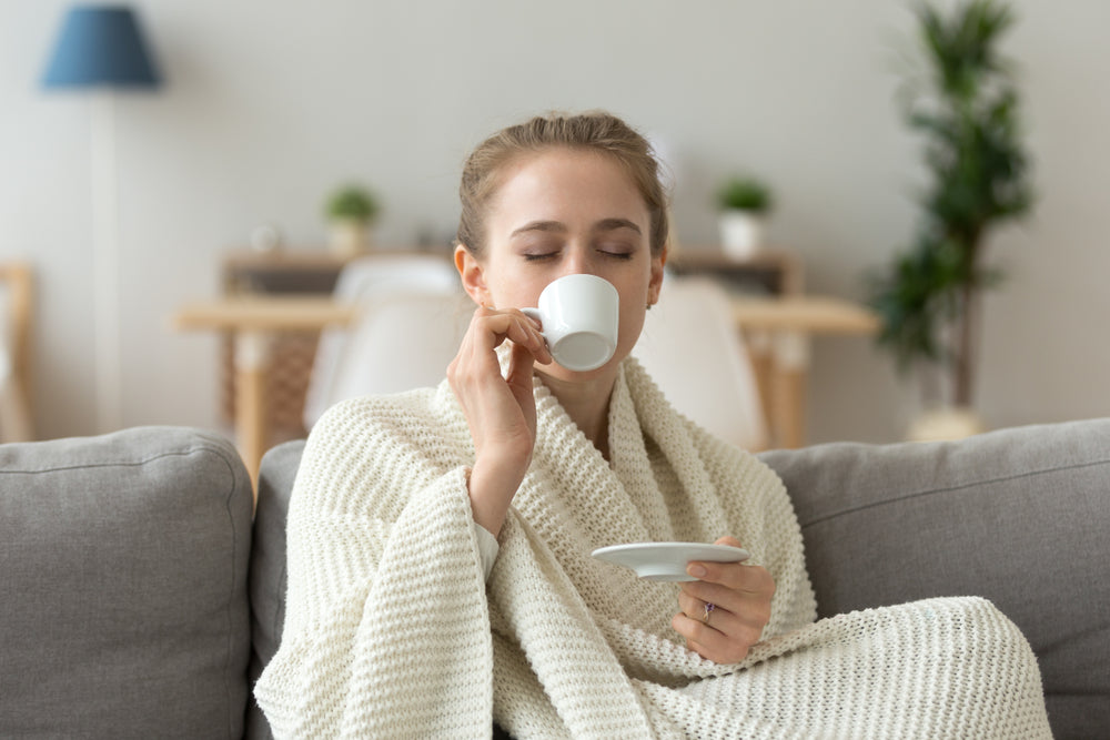 Happy female sit on couch in warm blanket enjoying cup of coffee