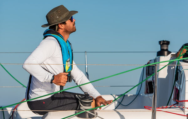 Ein Segler, der eine TeamO-Rettungsweste trägt, blickt auf die Segel der Yacht, während er das Focksegel mit einer Harken-Winde trimmt
