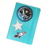 Spacelab 3 Embleem Met Space Shuttle Strijk Patch samen met een zilveren ster en astronaut patch op de voorkant van een blauwe agenda