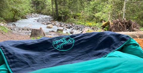 Camping bed set up outside in nature, with a river in the background. 