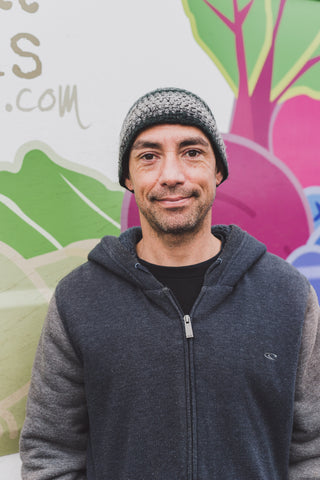 A white man is pictured from the waist up standing in front of an image of stylized veggies. He looks directly at the camera and is smiling slightly, and wearing a zip-up grey hoodie and grey beanie.