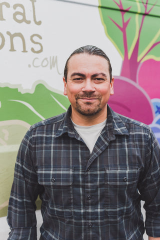A light-skinned person with dark hair smiles at the camera. He has a short beard and is pictured from the waist up, wearing a blue flannel and standing in front of a stylized illustration of veggies.