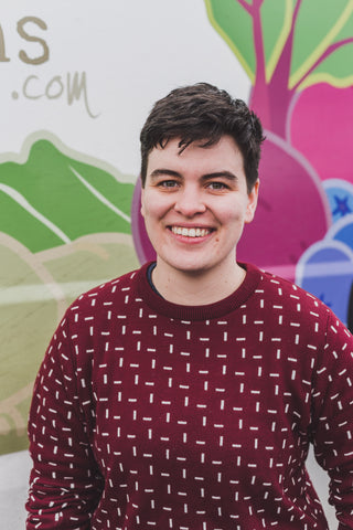 A white person with short brown hair smiles at the camera. They are pictured from the waist up, are wearing a maroon sweater with a cream geometric pattern, and are in front of a stylized image of veggies.