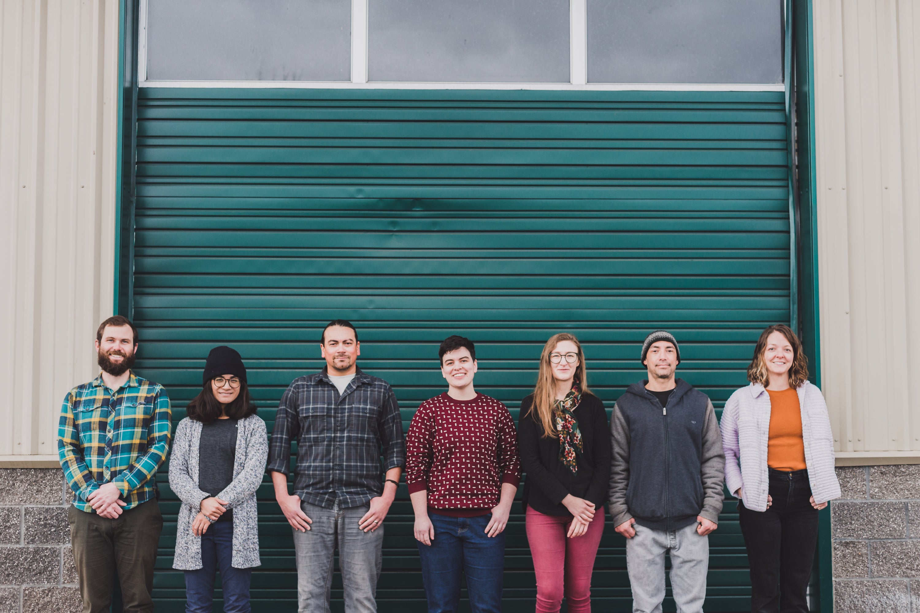 The 7 members of the spring 2023 Agricultural Connections Cooperative team stand in a line, facing the camera, in front of a green warehouse garage door. They wear a variety of sweaters and flannel with jeans and have their hands clasped or in their pockets. Left to right: Jake, Marisa, Miguel, Erin, Leslie, Josh, and Alex
