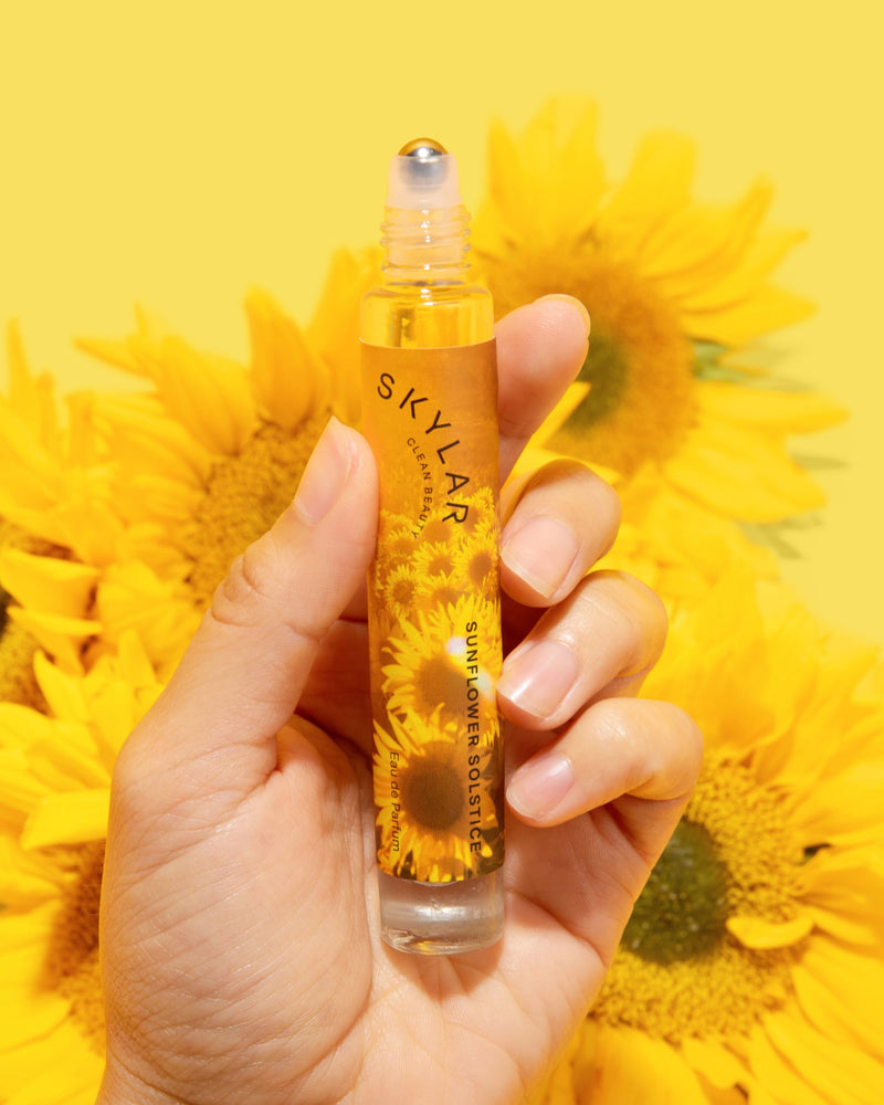 Woman holding Sunflower Solstice floral citrus scent in a travel-size rollerball above sunflowers.