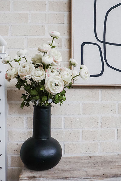 A faux flower arrangement of white and cream ranunculus sprays in a black vase.