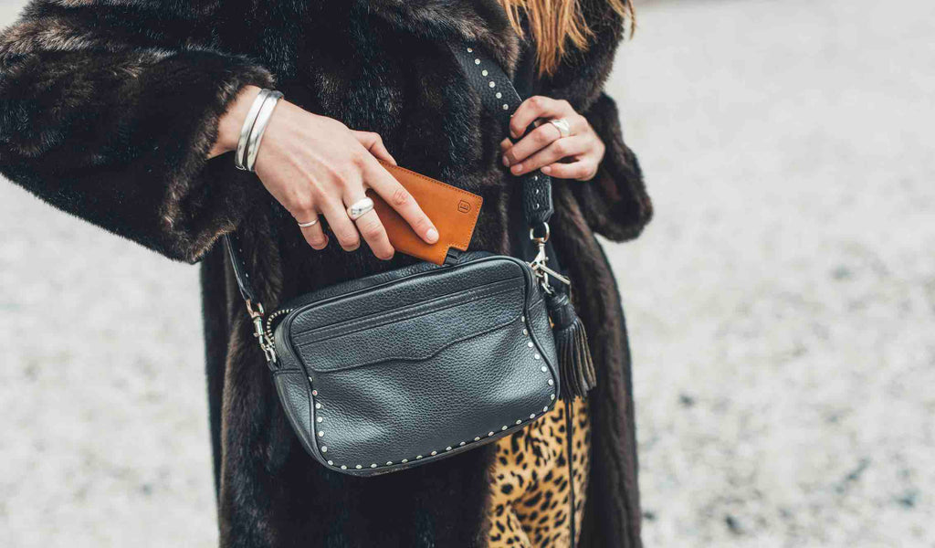 The image shows a woman dressed in a large faux fur coat, holding a black leather bag. She is sliding her Ekster Vachetta Parliament in Brescia Bronze into her wallet.