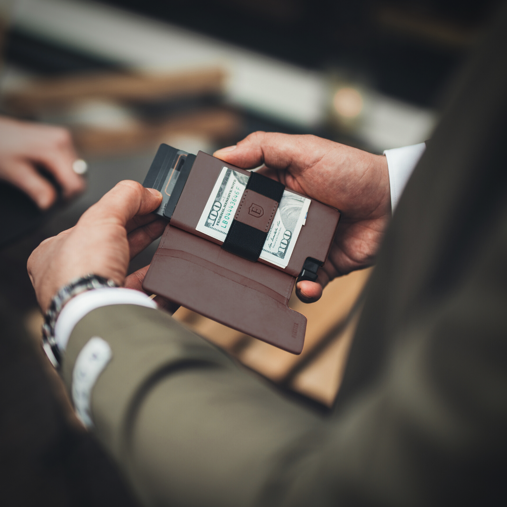 Pictured are a man's hands, in his right hand he is holding a brown Ekster Parliament Wallet and with his left hand he is removing a card from the wallet.