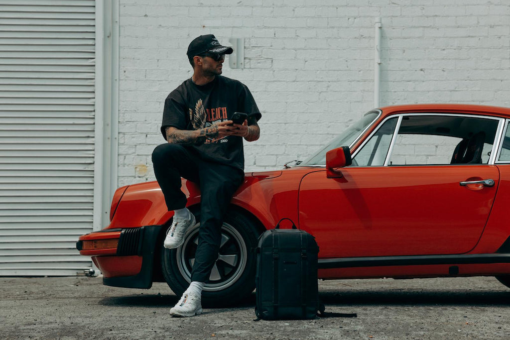 man leaning against red car with backpack