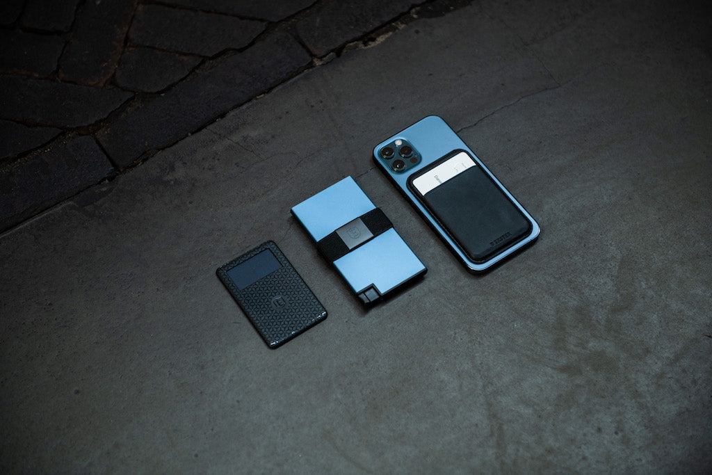 A tracker card, aluminum cardholder, and iPhone lying side by side on concrete. 