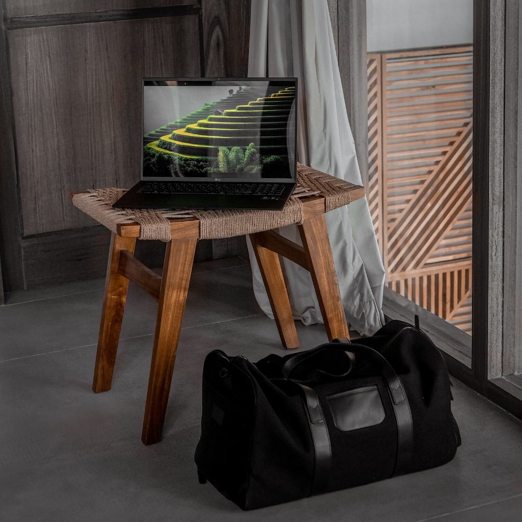 a weekender bag on the floor next to a table with an open macbook on it