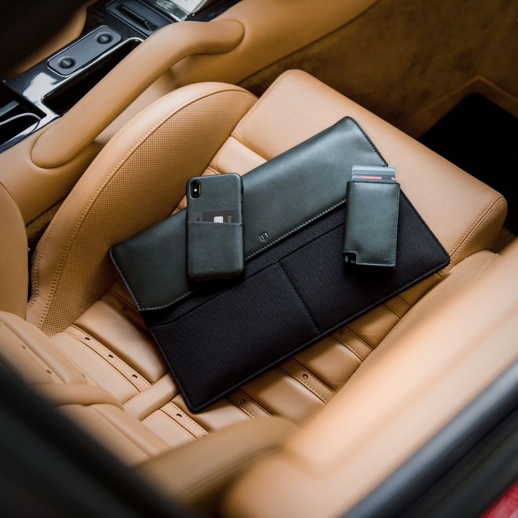 A black slim leather wallet, iPhone, and leather laptop sleeve on a luxury leather car seat.