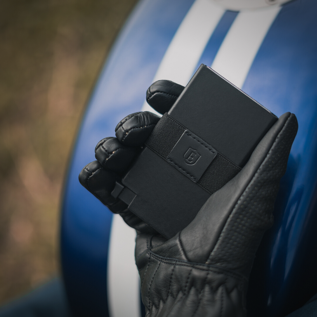 A man's hand wearing a leather glove holding a slim black leather cardholder wallet.