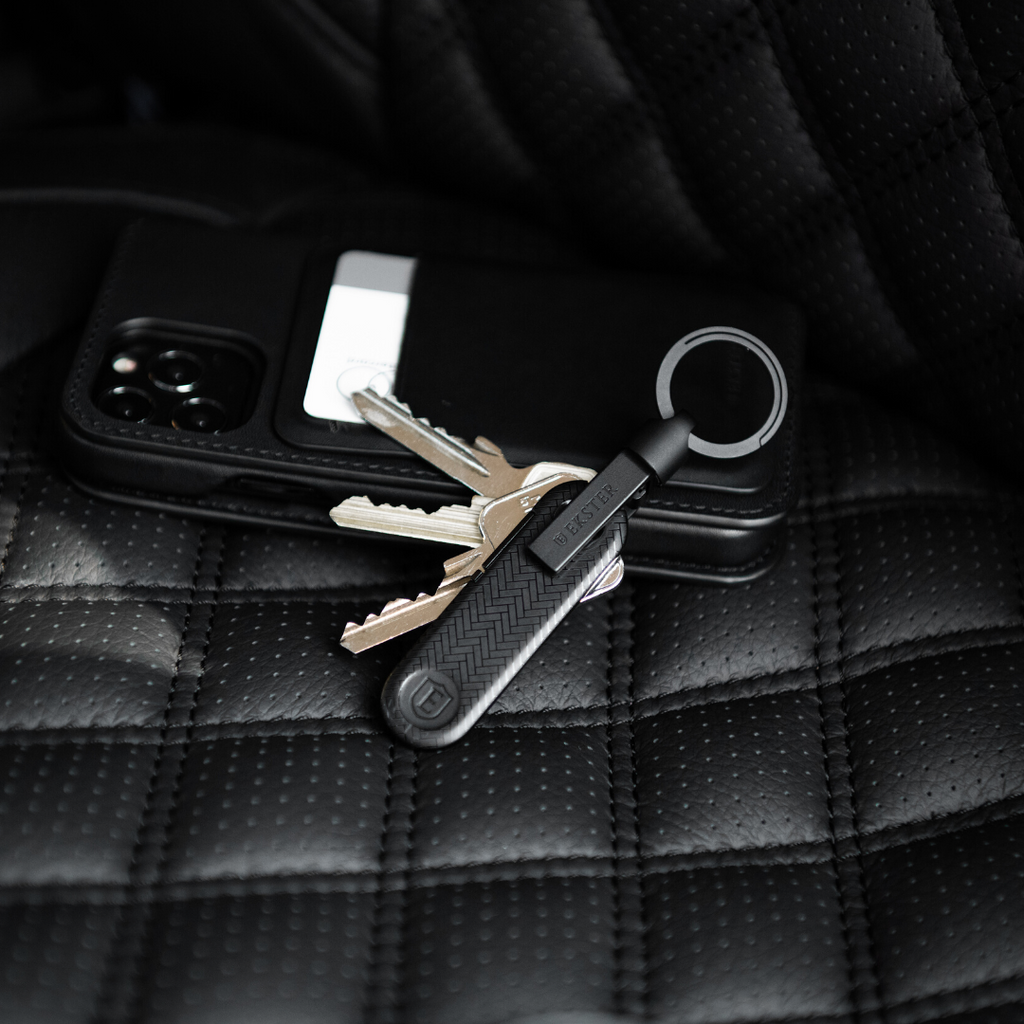 A close up picture of a black iPhone and an Ekster Key Holder and Tracker lying on a black leather car seat. 