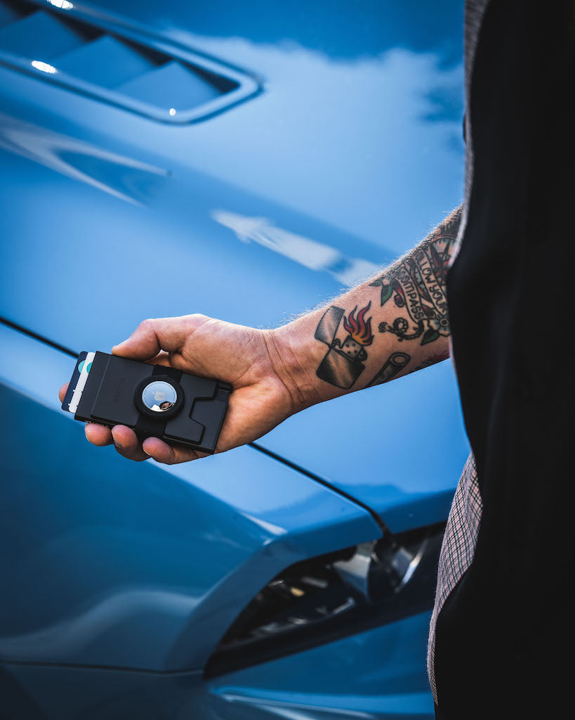 man's left hand holding a slim pop up wallet for airtag with the hood or a blue car in the background