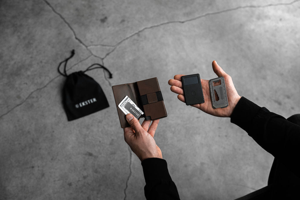 man's hands holding wallet, tracker card, and cash clip with grey industrial background