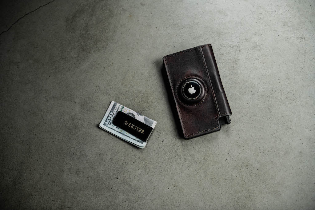 A black cash clip and a black airtag wallet are lying on a grey concrete surface