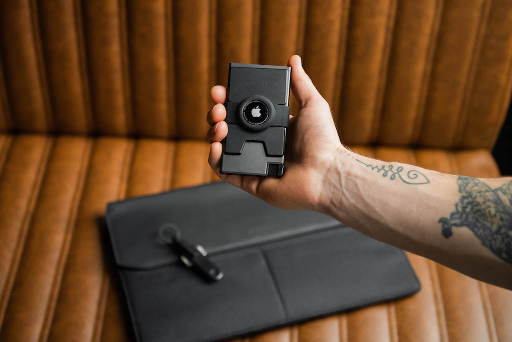 A man's hand is holding an Ekster black airtag cardholder wallet, in the background there is an Ekster black laptop sleeve and key tracker