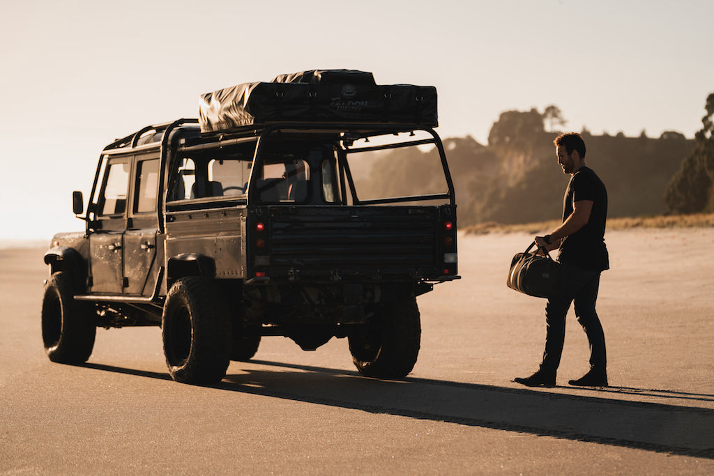 man holding weekender bag next to car on the beach