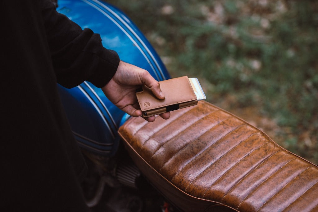 A man's left hand holding a slim leather wallet, in the background there is the leather seat of a motorcycle.