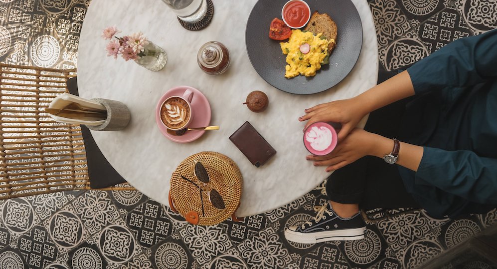 Top shot of a white table. A woman's hands are visible, holding a cup of coffee. The table is set with breakfast and another cup of coffee, as well as an Ekster trackable smart wallet, or slim wallet, and a pair of sunglasses. Some newspapers are also visible. 