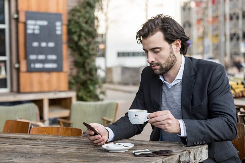 Image of a man in business attire drinking coffee, as he checks his iPhone. There is a travel security wallet on the table in front of him.