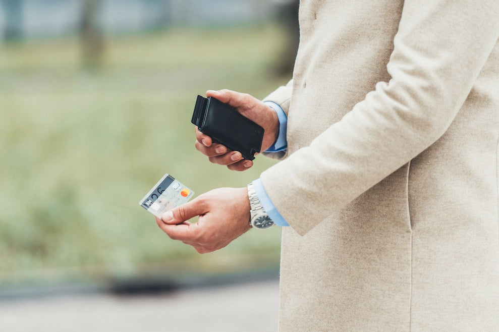 Image of a man's hands: one holds a black Ekster Parliament wallet, and the other hand has an N26 bank card in it. The wallet's card access feature shows all the cards displayed.