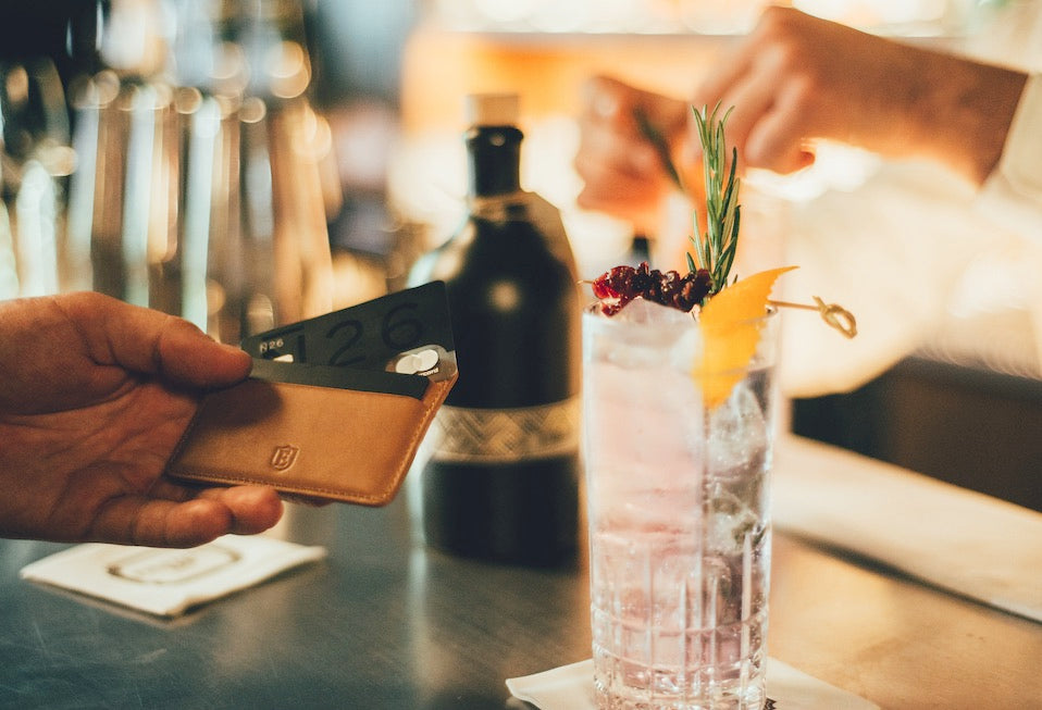 Image of a man's hand holding a Senate cardholder, with an N26 bank card peeking out of it. The Ekster logo is prominently displayed. Other elements of the picture include a bar as the background, with a cocktail on the side. 