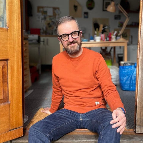 An image of James Green wearing an orange jumper and sitting in the open doorway of his printmaking studio.