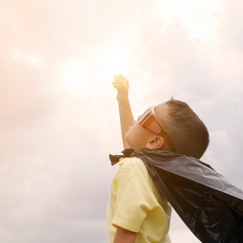 boy wearing a superhero cape with his arm stretched up to a sunny sky