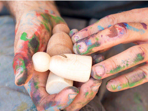 hands spalttered with colourful paint holding an unfinished wooden nin peg doll by Grapat toys