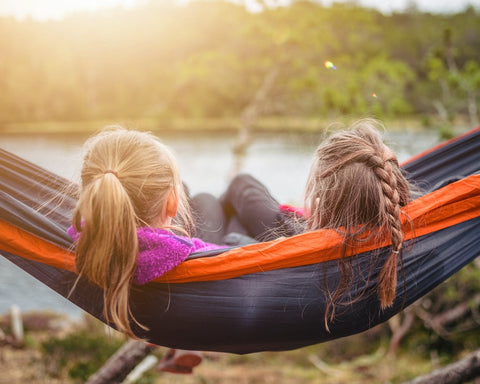 Two girls are relaxing in a hammock outside in front of a lake