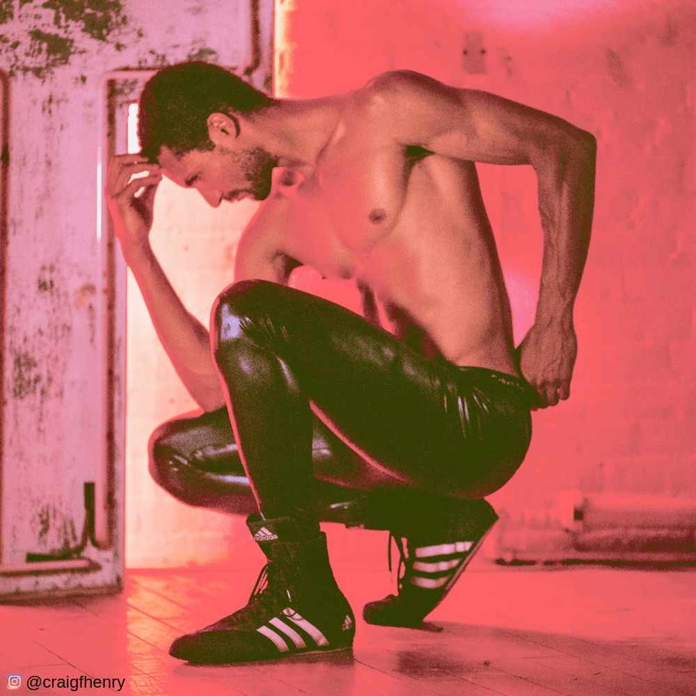 shirtless man with six pack kneeling in shiny black meggings red background