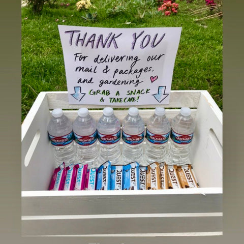 Bottles of water and Quest Protein Bars set out for delivery workers outside a home, with a sign that reads "Thank you for delivering our mail & packages. Grab a snack & take care!