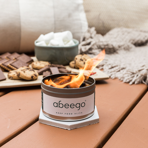 Abeego Mini Campfire is lit and burning, sitting on top of a white hexagon trivet with a board of smores ingredients in the background.