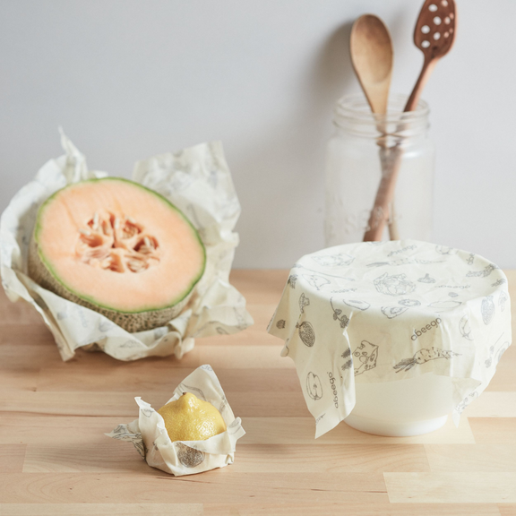 A small, medium, and large square Abeego food wrap covering a half lemon, half melon, and bowl.