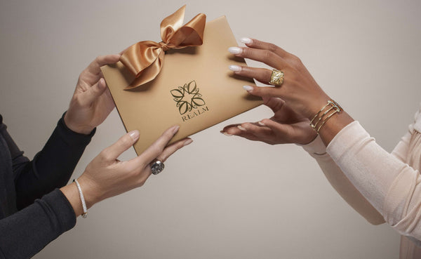 Two women wearing REALM Fine + Fashion Jewelry rings and bracelets pass each other a REALM signature gift bag with bow