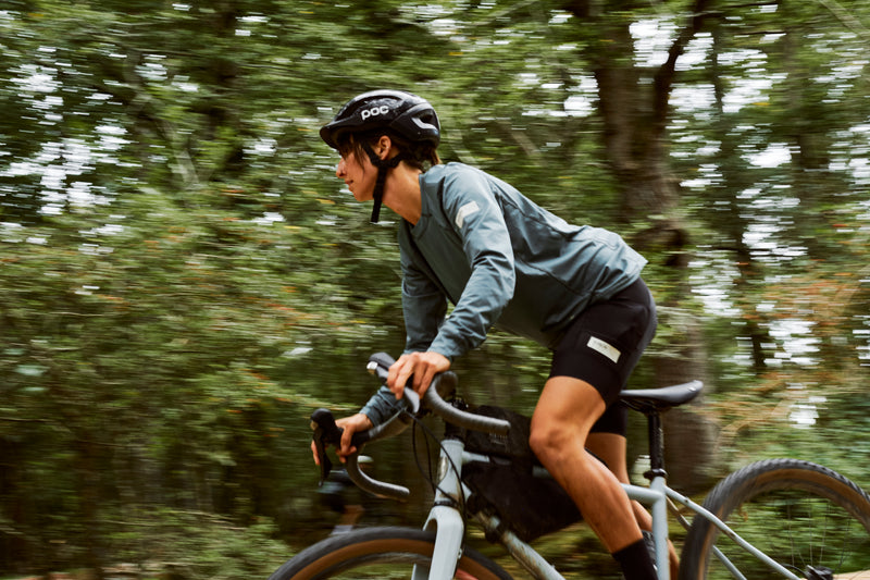 Woman riding a bike in the woods wearing a blue riding top and black cycling shorts