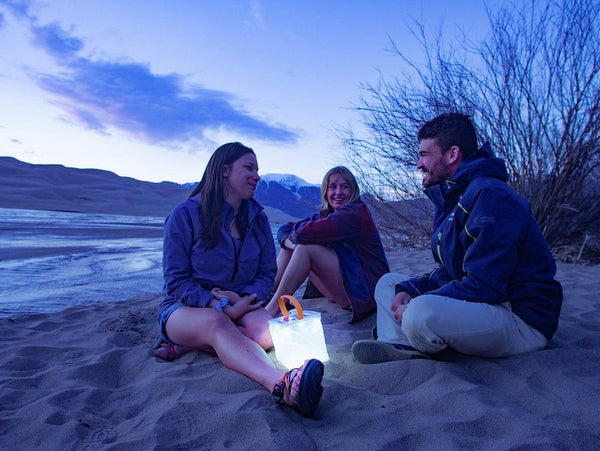 3 people siting on a beach with a LuminAID lantern